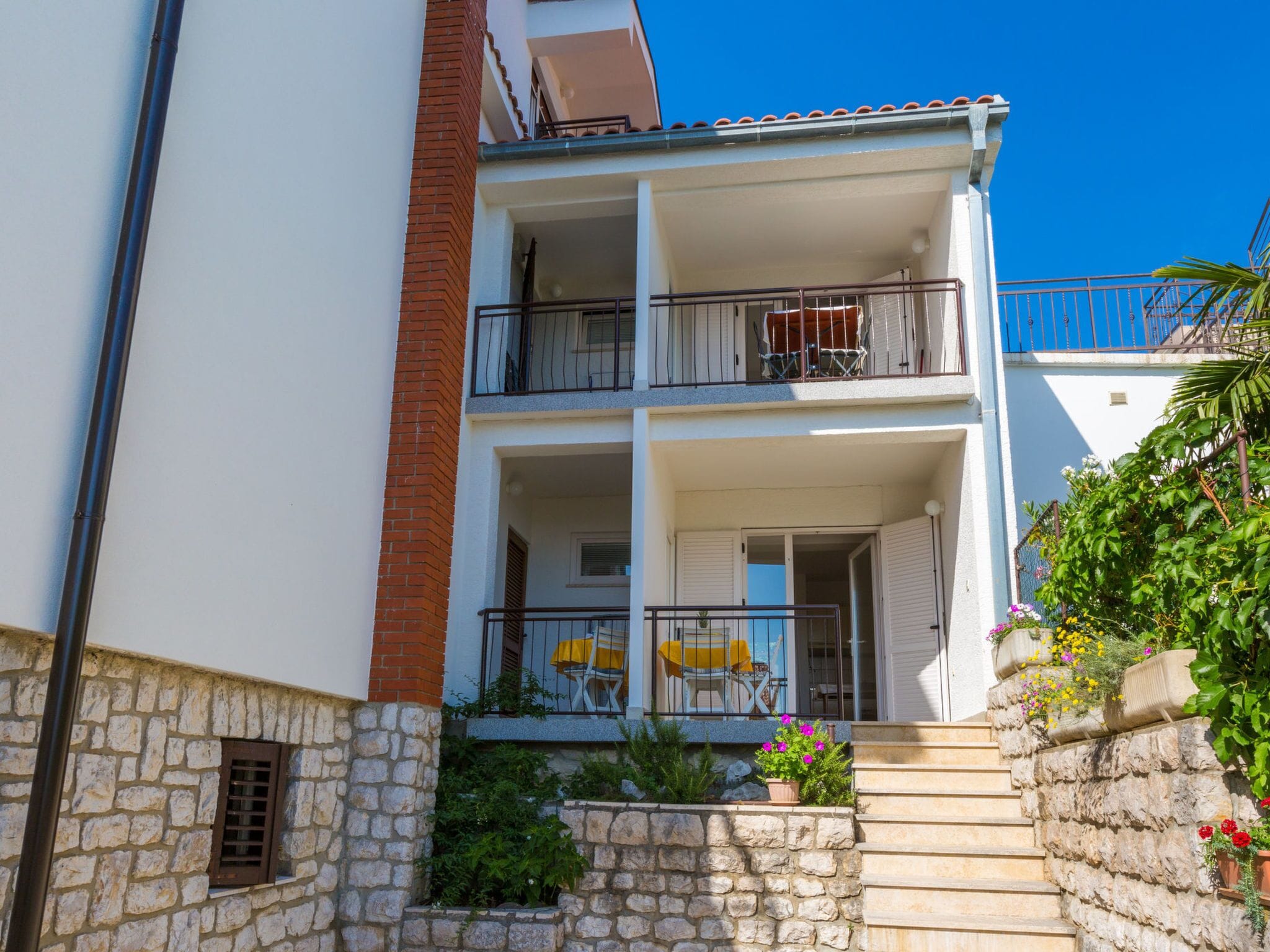 A two-story building with a stone facade, a balcony with a metal railing, and a small garden area with potted plants.