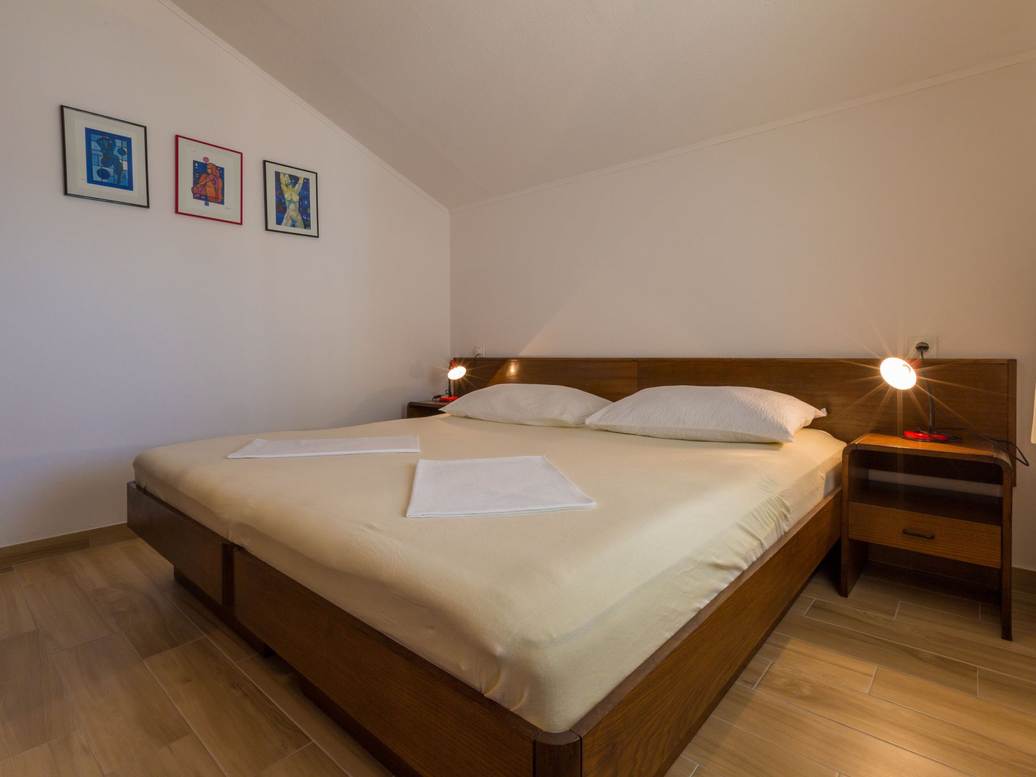 A neatly made bed with a wooden headboard, two bedside tables with lamps, and three framed artworks on the wall.