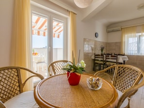 A dining area with wicker chairs, a round table with a woven mat, a red pot with flowers, and a buffet table with food items. - IRIS 3 / HR-02362-03 by Interhome (Crikvenica)