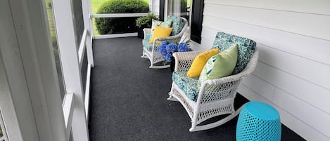 Eine überdachte Veranda mit drei Korbstühlen, jeder mit Kissen, und einem blauen gewebten Hocker.