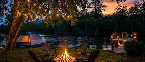 A campsite with a tent, a fire pit, and chairs by a lake at dusk.