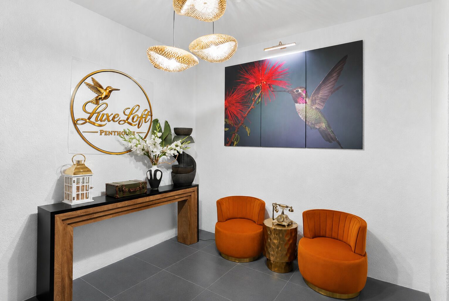 A reception area with a 'Live Loft' sign, a wooden console table with decorative items, two orange chairs, and a framed bird painting.