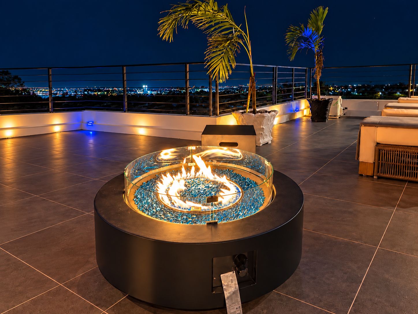A rooftop terrace with a circular fire feature surrounded by blue glass pebbles, palm trees, and a cityscape view.