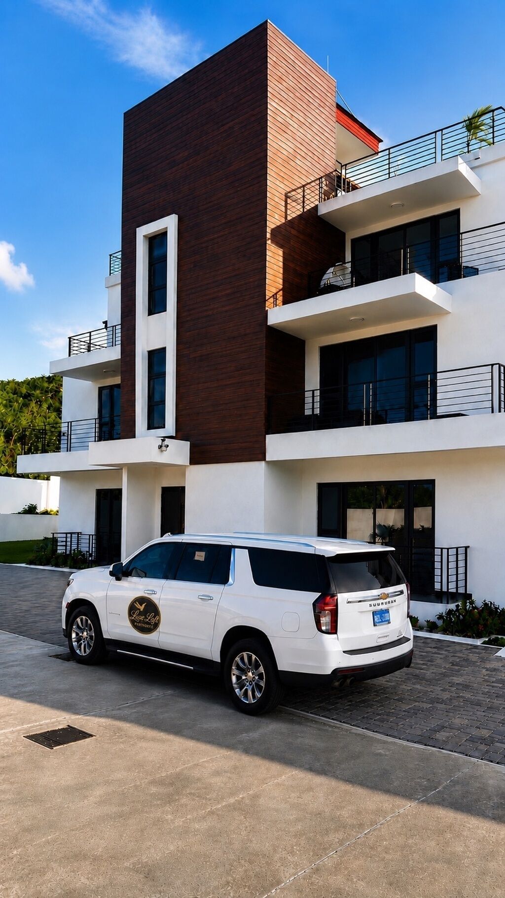 A modern multi-story building with a white and brown facade, a white SUV parked in front, and a clear blue sky.
