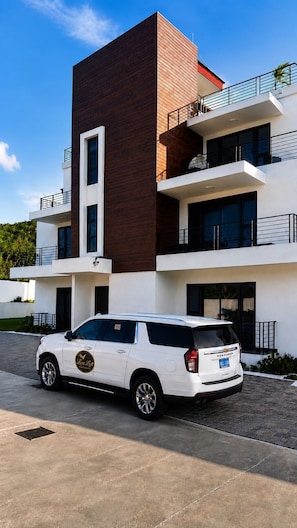 A modern multi-story building with a white and brown facade, a white SUV parked in front, and a clear blue sky. - Luxe Loft Penthouse, a luxury property in Montego Bay, 6 mins from MBJ airport (Montego Bay)