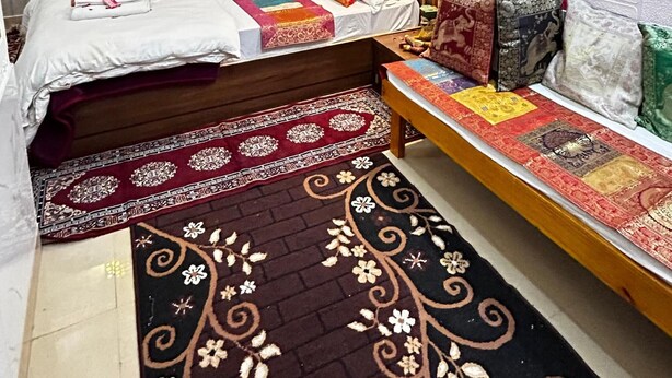 A bedroom with a bed, a bedside table, a wall-mounted lamp, a window with curtains, and a decorative rug. - Hotel casa del sol (Jaisalmer)