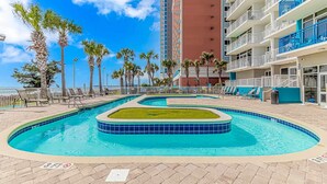 A pool area with multiple swimming pools, palm trees, and a tall building in the background. - Atlantica 452 Oceanfront Escape (Myrtle Beach)