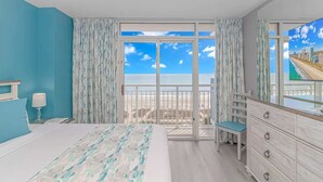 A bedroom with a bed, a dresser, a chair, and a large window with a view of the beach. - Atlantica 452 Oceanfront Escape (Myrtle Beach)