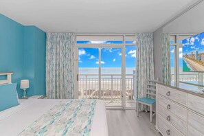 A bedroom with a bed, a dresser, a chair, and a large window with a view of the beach. - Atlantica 452 Oceanfront Escape (Myrtle Beach)