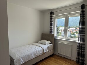 Una habitación pequeña con cama individual, piso de madera, ventana con cortinas y vistas a edificios y montañas.