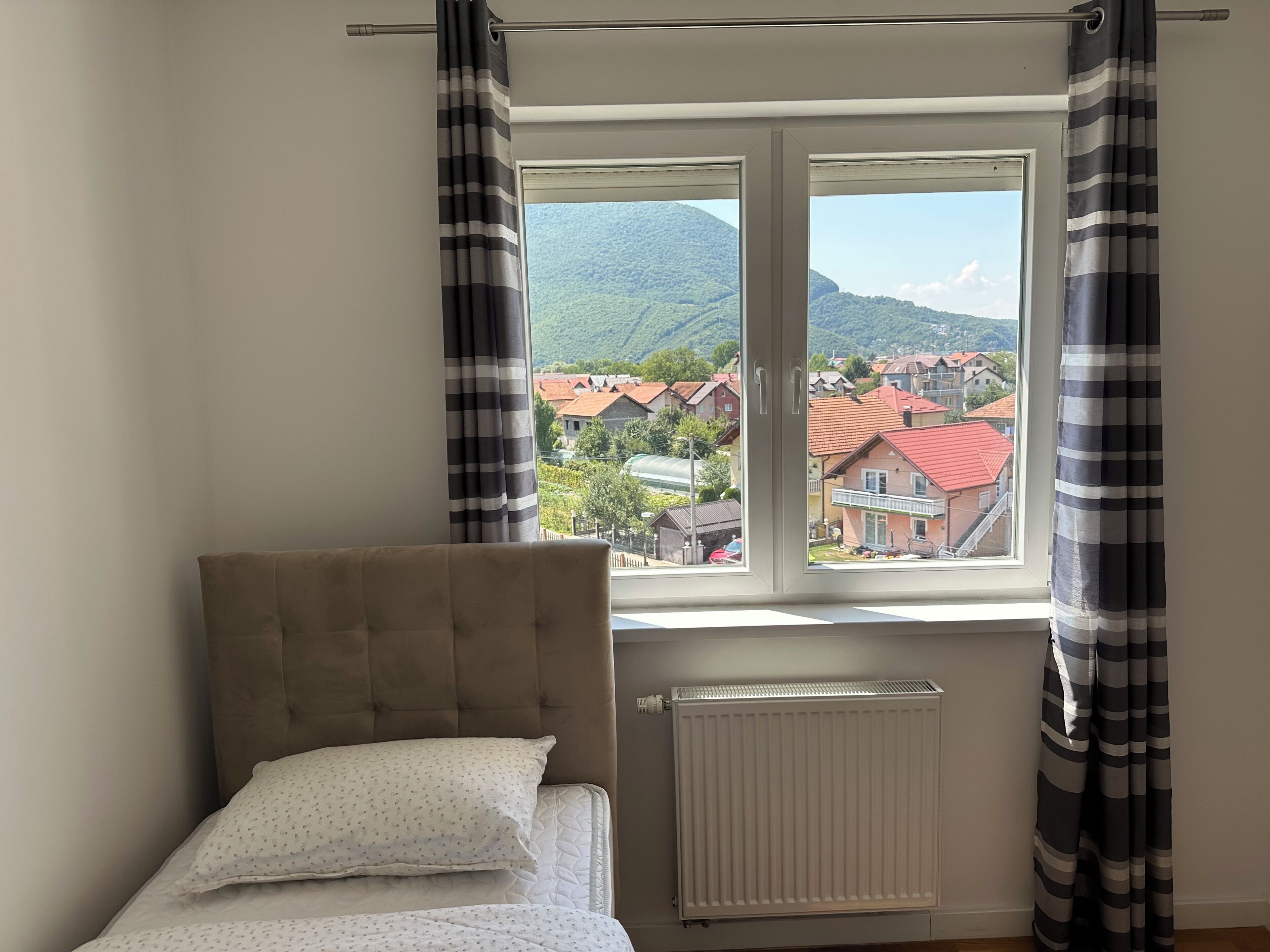 A bedroom with a bed, a window with curtains, and a view of houses and mountains.