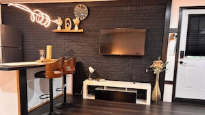Een moderne keuken met een donkere bakstenen muur, een wit aanrecht, twee barkrukken, een televisie en een deur.