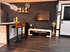 Une cuisine moderne avec un mur en briques sombres, un comptoir blanc, deux tabourets de bar, un téléviseur et une porte.