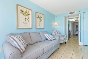 A living room with a grey sofa, striped armchair, and a lamp. There are framed botanical prints on the wall. - Oceanfront 1BR Retreat for 4! Atlantica 200 (Myrtle Beach)