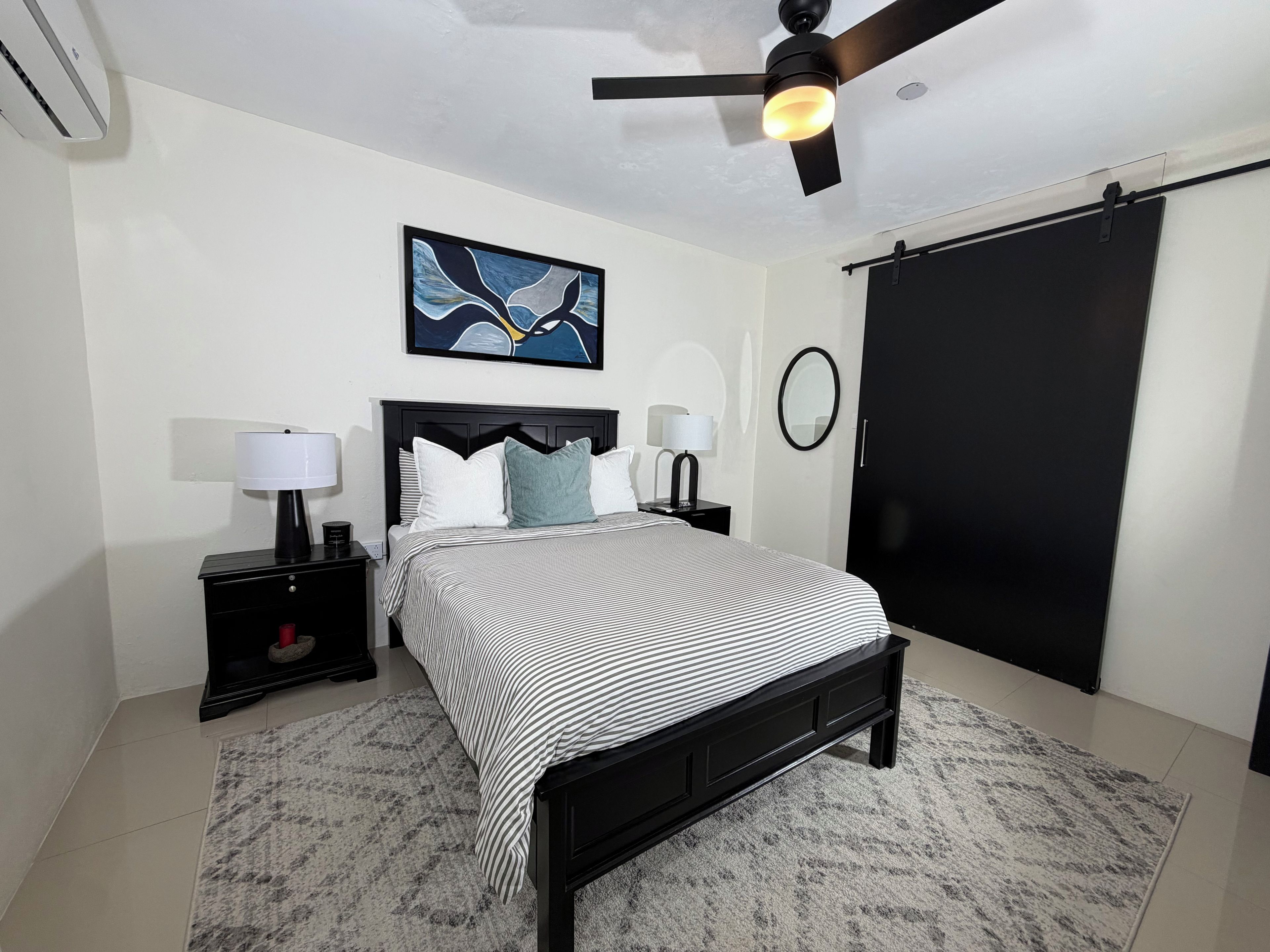 Une chambre à coucher comprenant un lit, deux tables de chevet, un ventilateur de plafond, une œuvre d’art accrochée au mur, un miroir et une armoire noire.