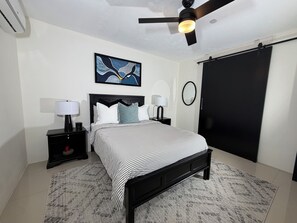 Une chambre à coucher comprenant un lit, deux tables de chevet, un ventilateur de plafond, une œuvre d’art accrochée au mur, un miroir et une armoire noire.