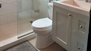 Et moderne badeværelse med en glasafskærmet bruseniche, et hvidt toilet, en vask med spejl og en hylde med toiletartikler.