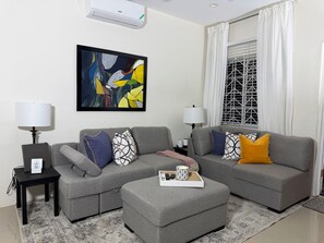 A living room with a grey sofa set, two side tables with lamps, a framed abstract painting, and a window with curtains. - Modern 1BR Getaway near Beaches, Airport, and popular entertainment spots. (Montego Bay)
