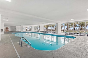 A large indoor swimming pool with clear glass walls, lounge chairs, and a view of palm trees. - Boardwalk 135 Oceanfront Getaway (Myrtle Beach)