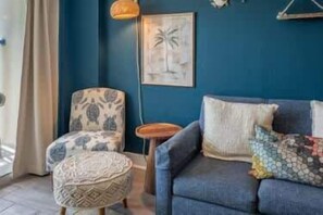 Ein Zimmer mit blauer Wand, einem gemusterten Sessel, einer Couch mit Kissen, einem kleinen runden Tisch und einer Pendelleuchte.