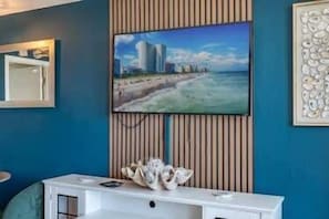 Ein Zimmer mit an der Wand befestigtem Fernseher, einem weißen Konsolentisch mit Dekoration und einem Bilderrahmen mit Strandmotiv.