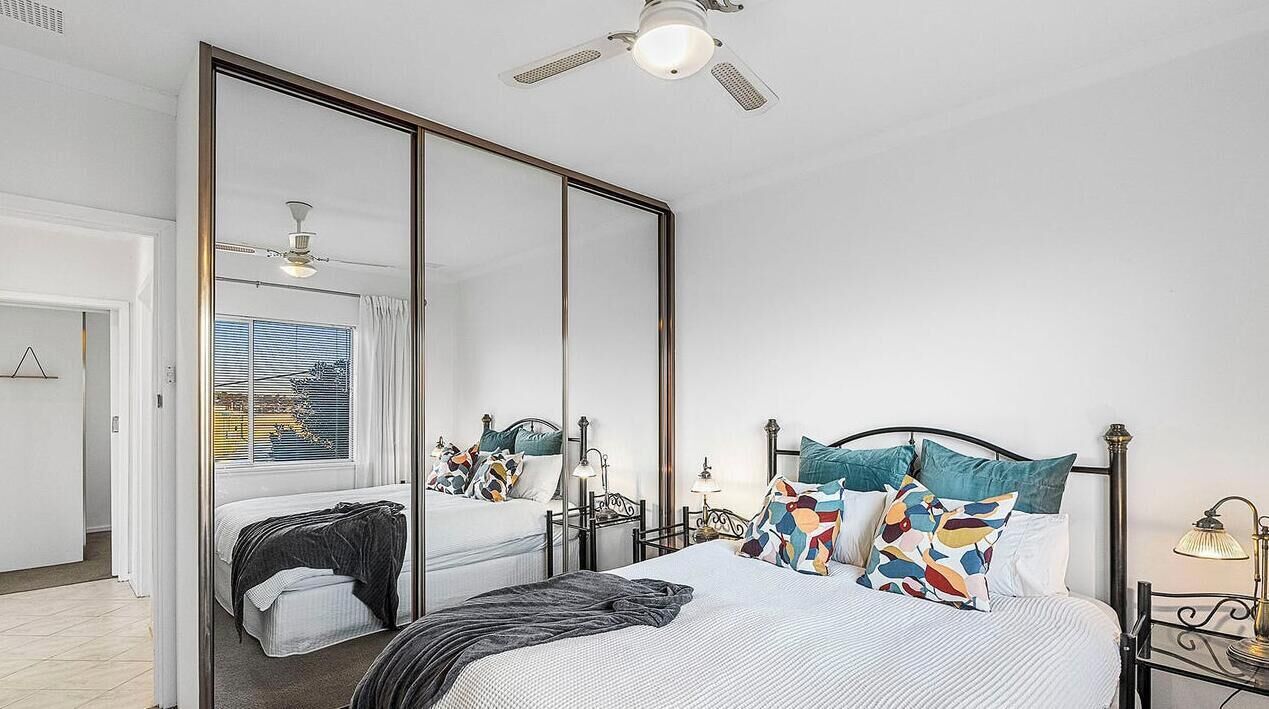 A bedroom with a large bed, a ceiling fan, a window with blinds, and a mirror.