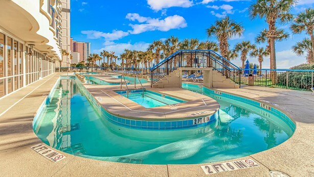 A pool area with multiple swimming pools, palm trees, and a bridge. - Boardwalk 440 Studio (Myrtle Beach)