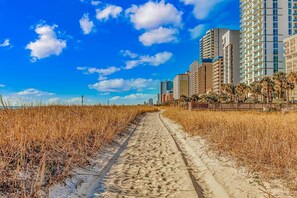 Ein sandiger Pfad führt zu einem Strand mit hoch aufragenden Gebäuden im Hintergrund.