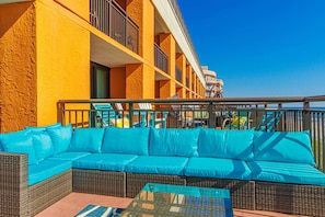 A balcony with a wicker sofa and chairs, a glass table, and a view of a building with balconies. - Caravelle 515 Studio (Myrtle Beach)
