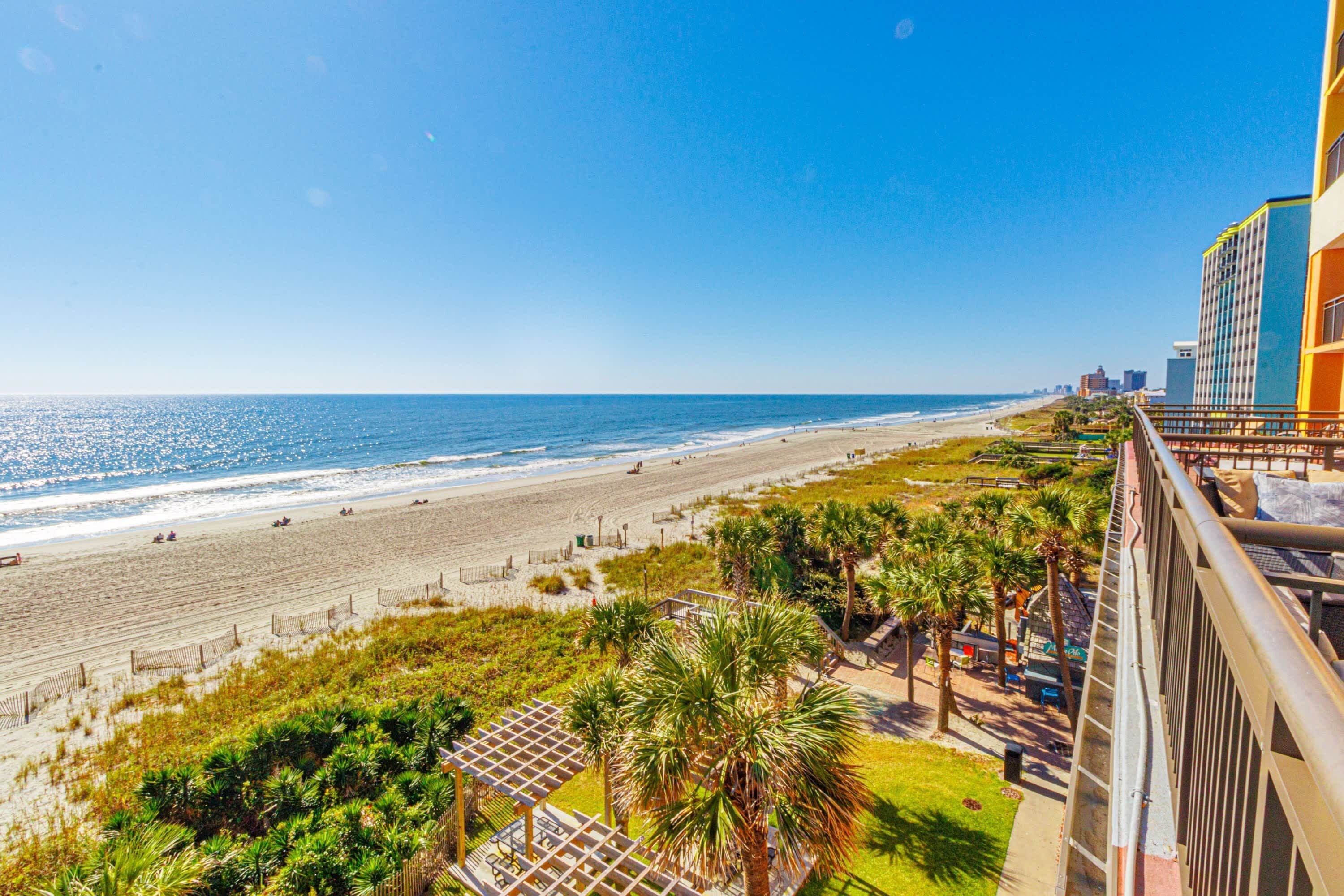 A beach view from a balcony overlooking a sandy beach with palm trees and a clear blue sky.
