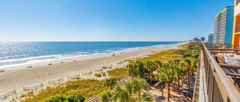 A beach view from a balcony overlooking a sandy beach with palm trees and a clear blue sky.