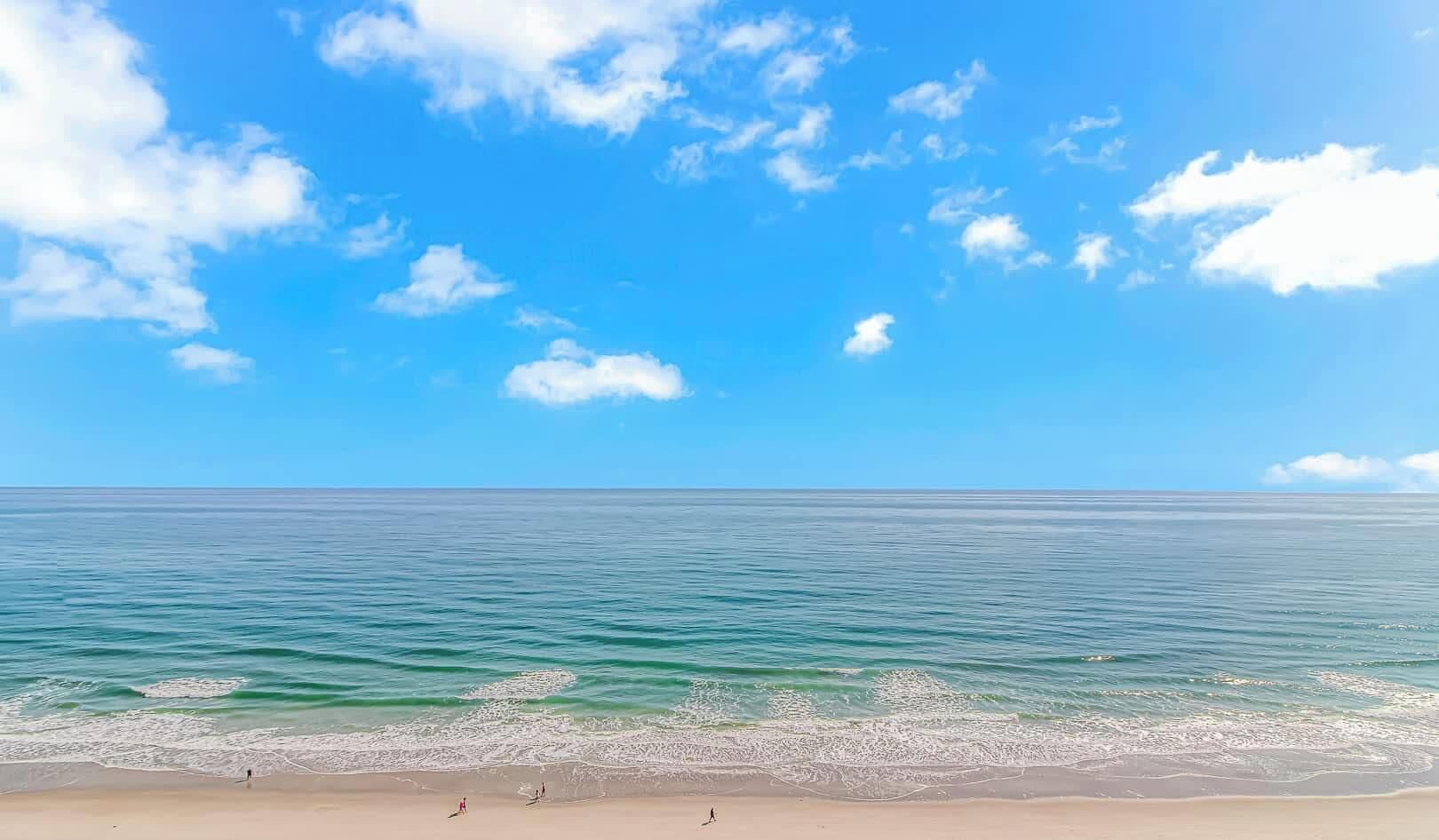A beach with clear blue water, a sandy shore, and a few people walking.