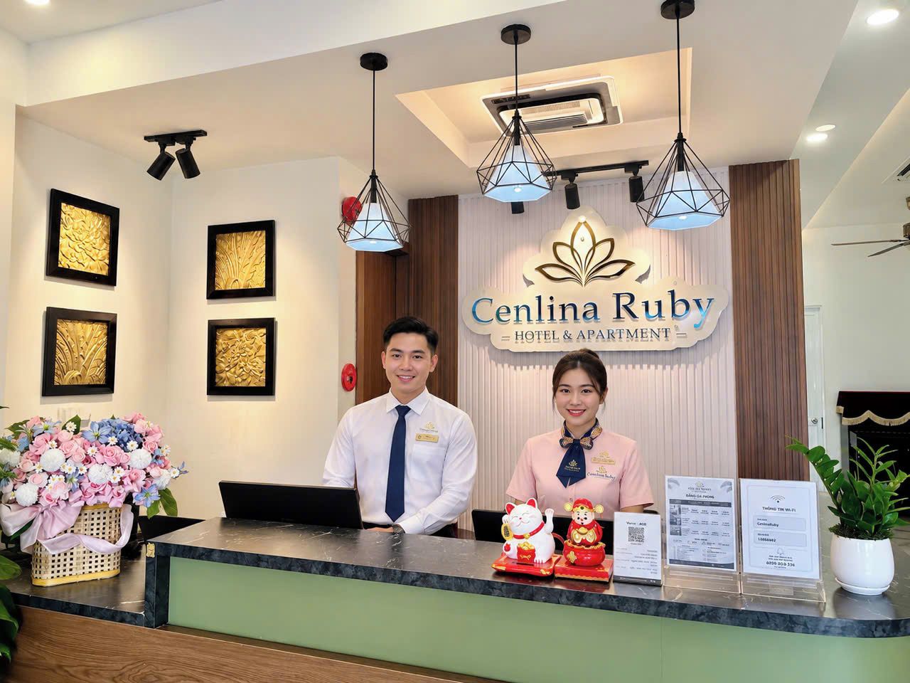 A hotel reception desk with framed artwork on the walls, a reception desk with a computer, and a sign that reads 'Cenlina Ruby Hotel & Apartment'.