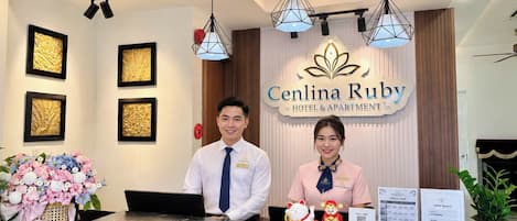 Balcão de recepção de hotel com obras de arte emolduradas nas paredes, um balcão com computador e uma placa com a inscrição 'Cenlina Ruby Hotel & Apartment'.