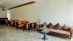 Un vestíbul d'hotel amb zona de seients que inclou cadires taronges i un sofà marró, una màquina de cafè i una planta decorativa.