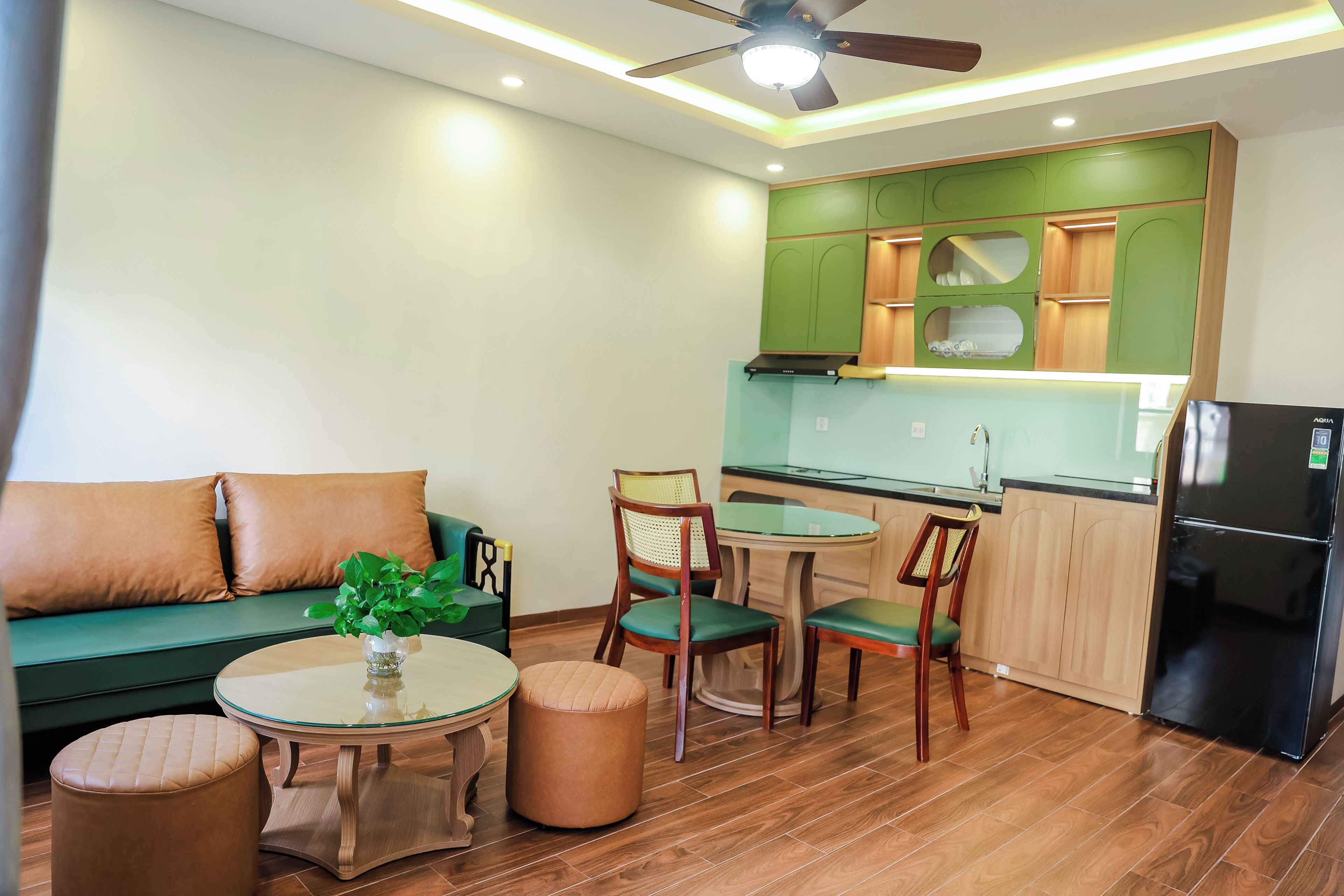 Un espace de vie compact comprenant un canapé, deux fauteuils, une petite table, un coin cuisine avec des meubles verts et un réfrigérateur noir.