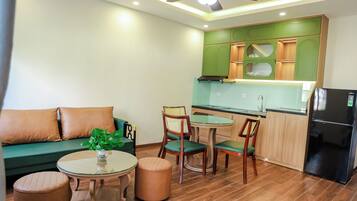 Un espace de vie compact comprenant un canapé, deux fauteuils, une petite table, un coin cuisine avec des meubles verts et un réfrigérateur noir.