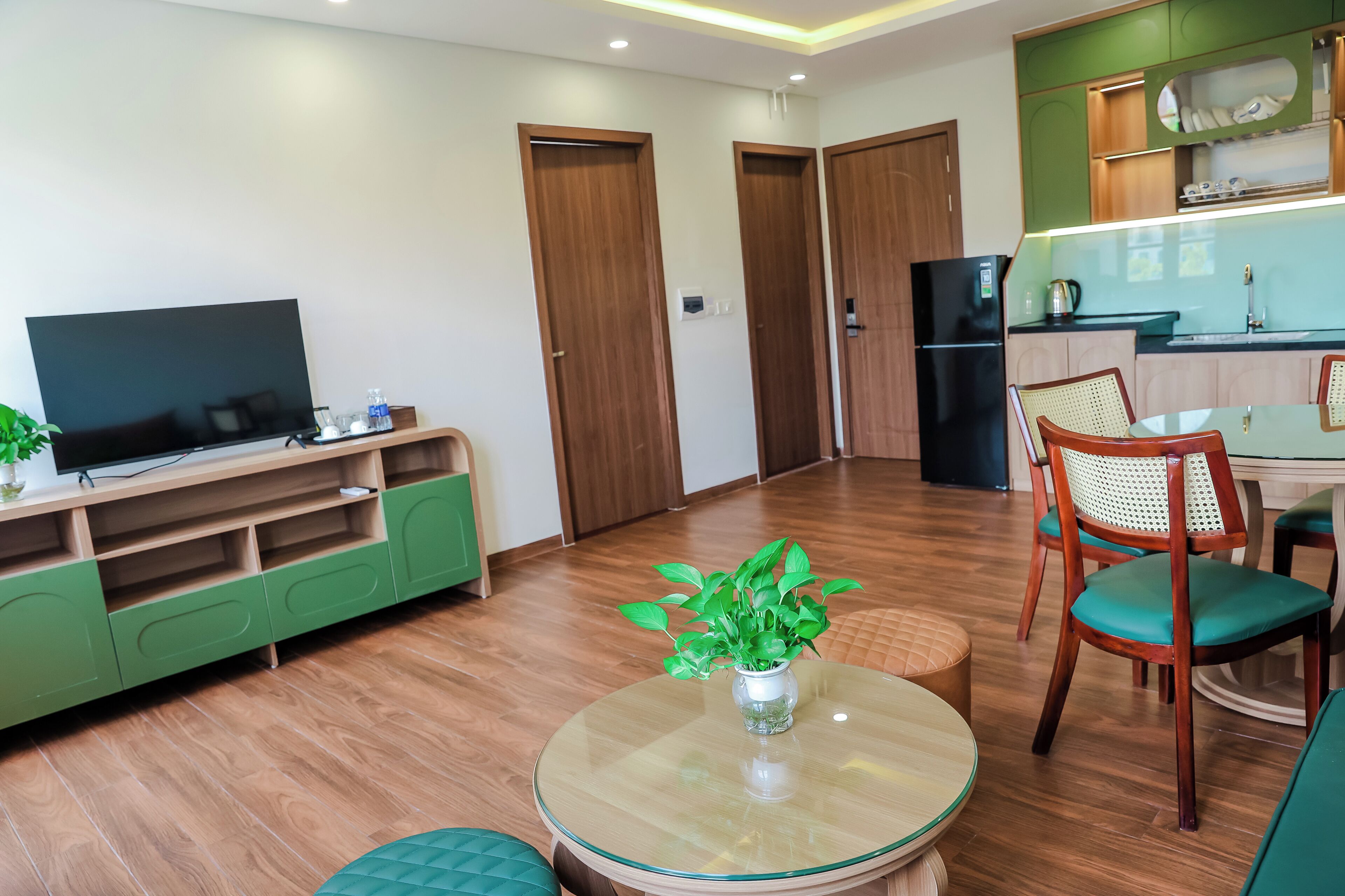 A modern living room with a flat-screen TV, green entertainment unit, and a dining area with a round glass table and chairs.