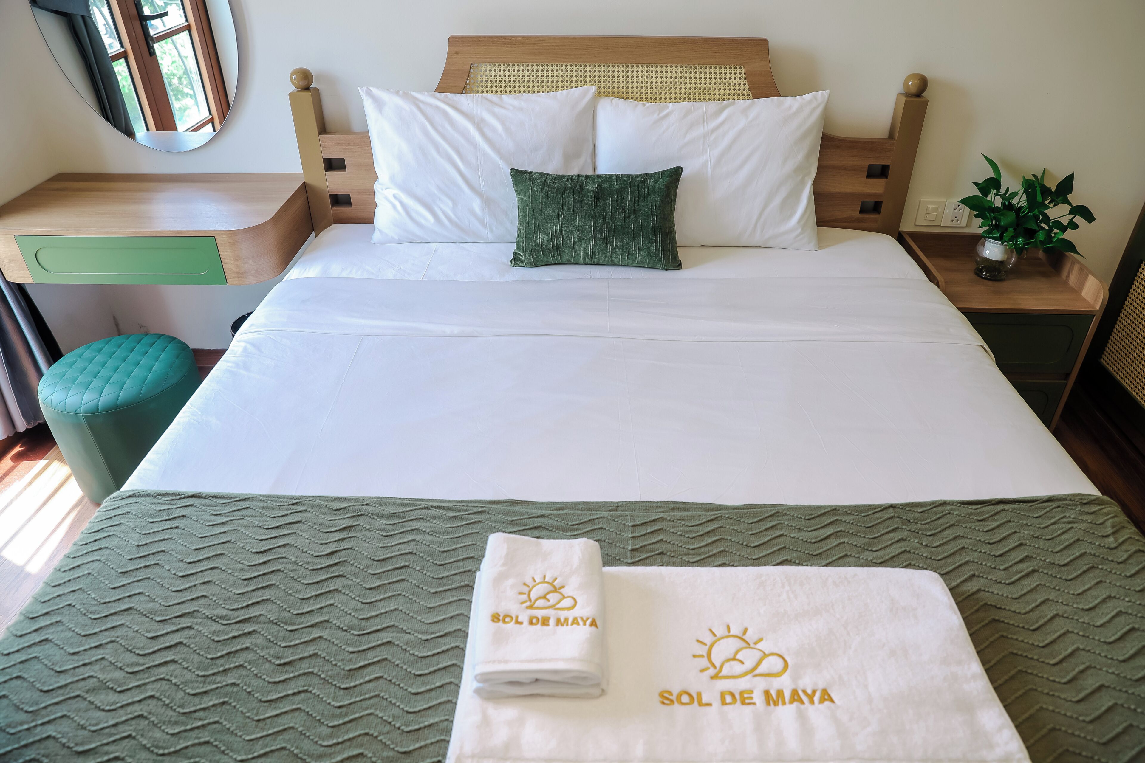 Una cama bien hecha con sábanas blancas, una almohada verde y un plaid verde, además de una toalla con el texto 'SOL DE MAYA'.