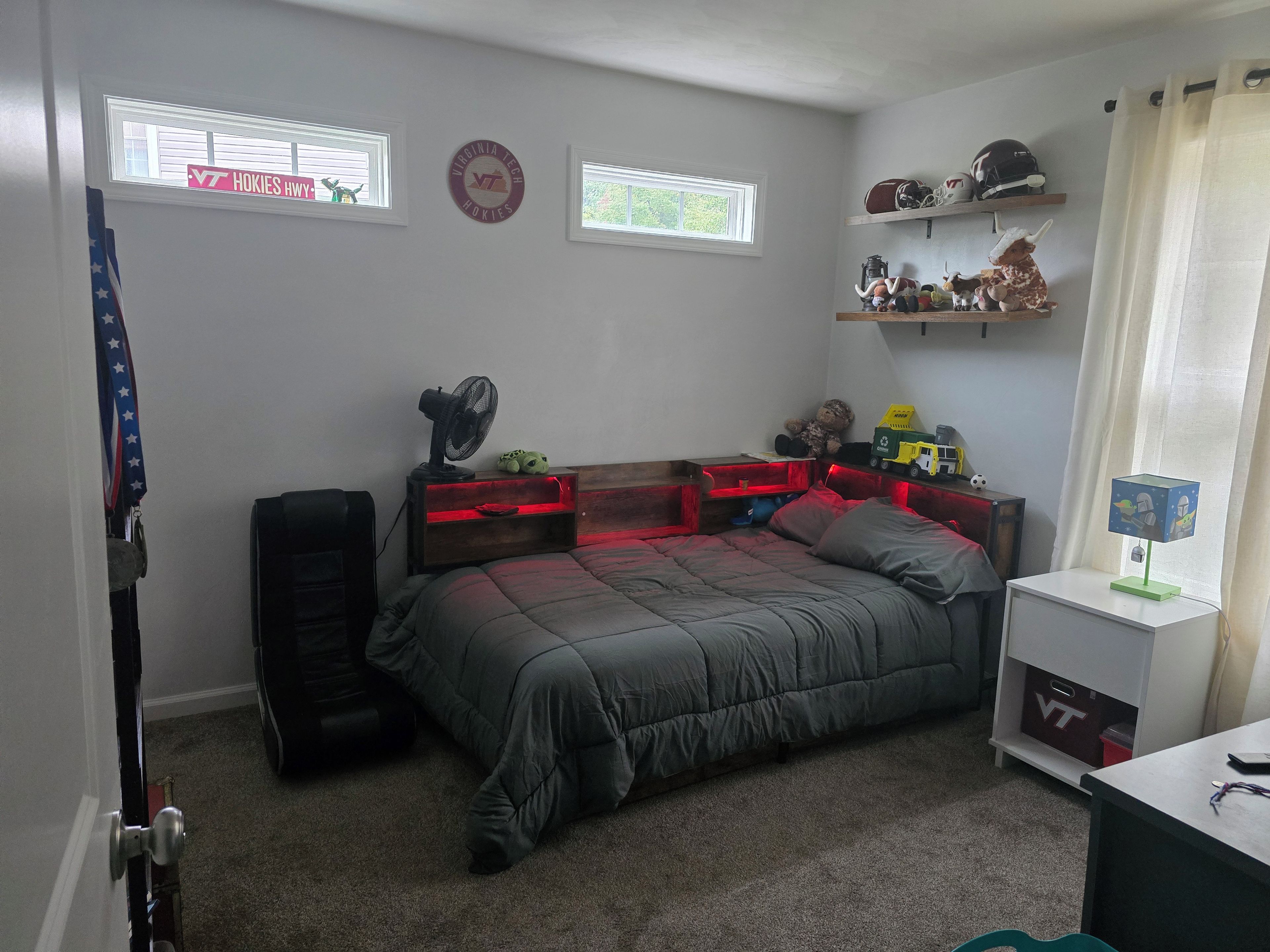 A bedroom with a bed, a fan, a shelf with helmets, and a window with a sign.