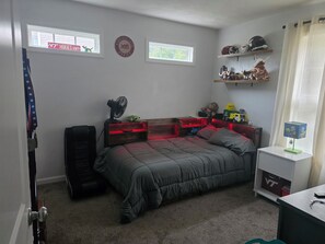 Een slaapkamer met een bed, een ventilator, een plank met helmen en een raam met een bord.