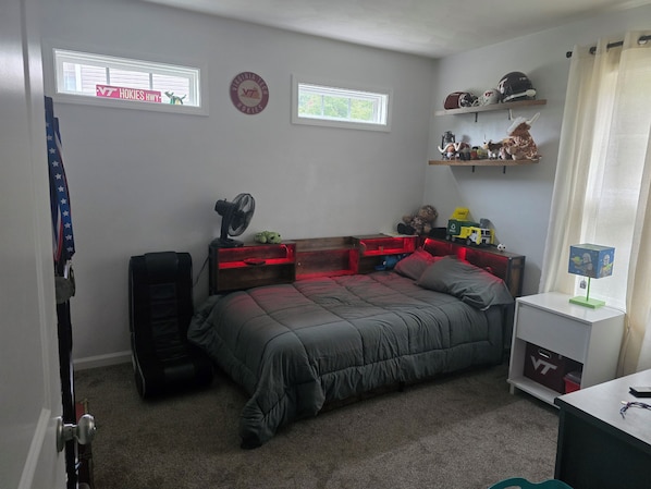 A bedroom with a bed, a fan, a shelf with helmets, and a window with a sign.