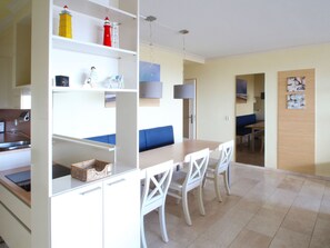A modern kitchen with white cabinetry, a dining area with a wooden table and white chairs, and a blue sofa. - Albatros by Interhome (Noordwijk)