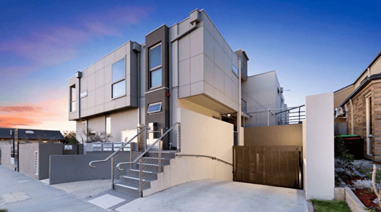 A modern two-story building with a staircase leading to the entrance, a gated driveway, and a clear sky in the background.