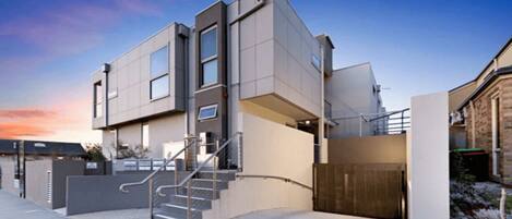 A modern two-story building with a staircase leading to the entrance, a gated driveway, and a clear sky in the background.