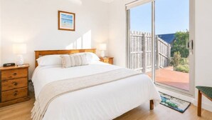 A bedroom with a bed, bedside tables, a nightstand, a window with a view of the outdoors, and a framed picture on the wall. - Bonbeach Retreat by Ready Set Stay - 2 Bedroom, Family Friendly Apartment (Bonbeach)