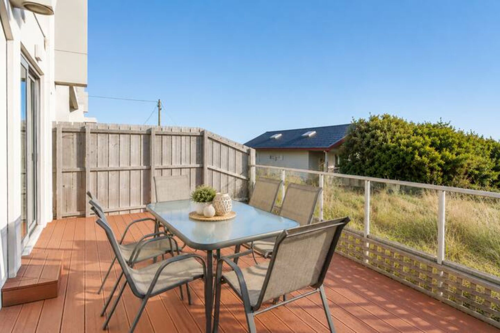 Een terras buiten met een tafel en stoelen, een houten omheining en uitzicht op een huis en groen.
