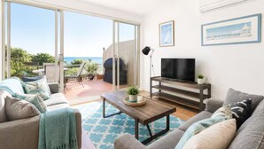 A living room with a sofa, a coffee table, a TV, and a balcony with a view of the sea. - Bonbeach Retreat by Ready Set Stay - 2 Bedroom, Family Friendly Apartment (Bonbeach)