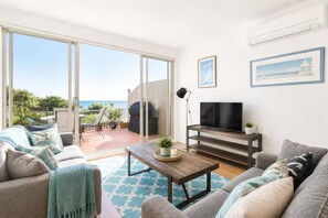 Een woonkamer met een bank, een salontafel, een televisie en een balkon met uitzicht op zee.