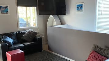 A living room with a black leather sofa, a red ottoman, a white coffee table, a flat-screen TV mounted on the wall, and a window with blinds.
